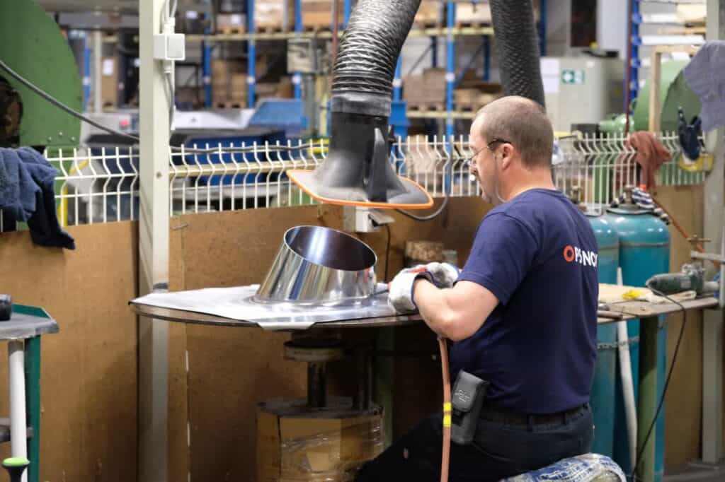 A factory worker wearing a navy blue shirt and protective gloves operates machinery in an industrial setting. He works on a shiny, metallic piece under a large, flexible exhaust hood. Behind him are various industrial equipment and tools.
