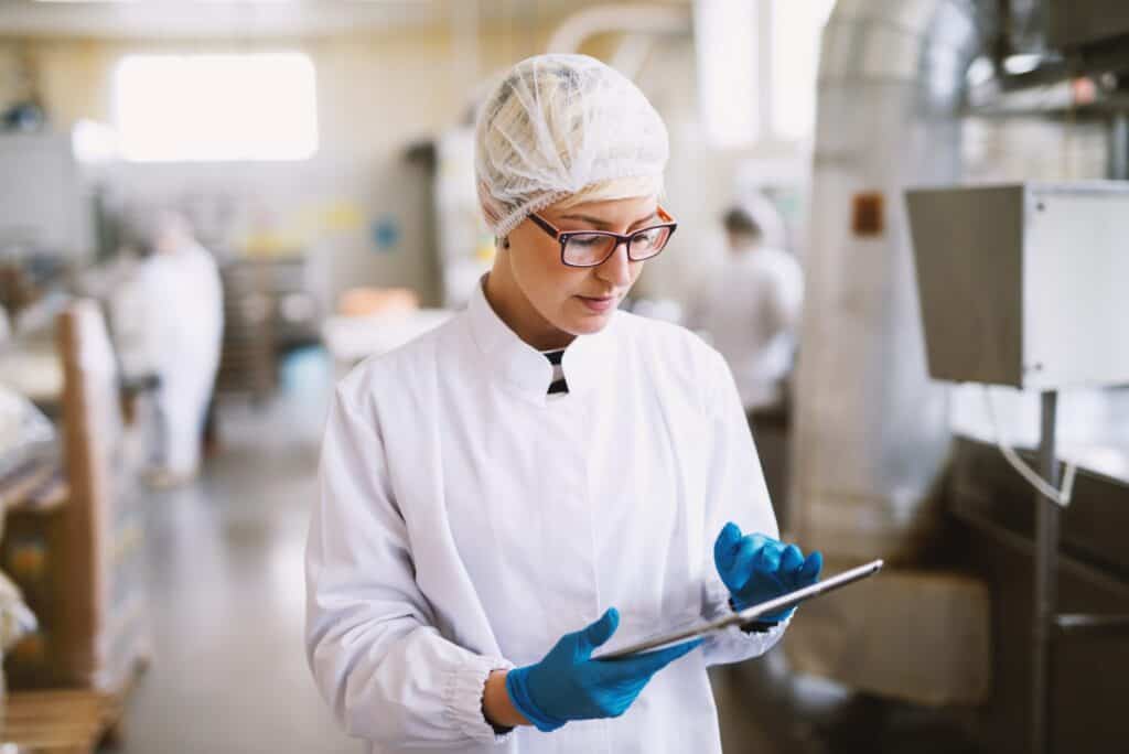 A person in protective gear, including a hairnet, eyeglasses, gloves, and a white lab coat, is using a tablet in a laboratory or industrial setting. The background shows equipment and a blurred figure of another worker.
