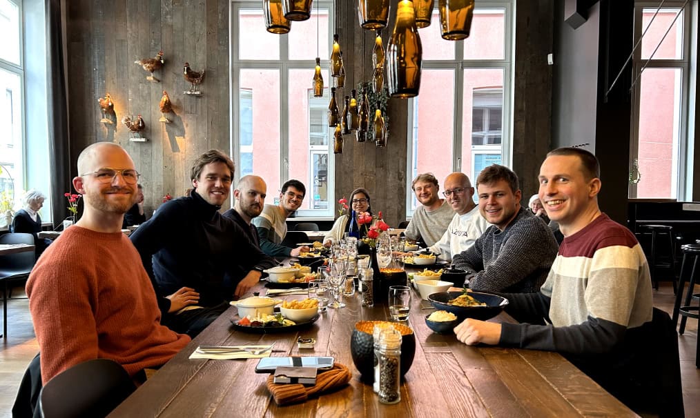 A group of ten people sits around a long wooden table in a restaurant, smiling at the camera. The table is set with glasses, dishes of food, and decorative items. Large windows in the background allow natural light to fill the space, highlighting the modern decor.