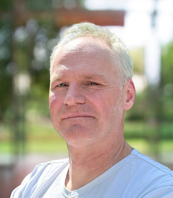 Elderly man with grey hair outdoors, sunlight highlighting his face, casual white T-shirt, natural background, expression of confidence and experience, symbolizing wisdom and life journey.