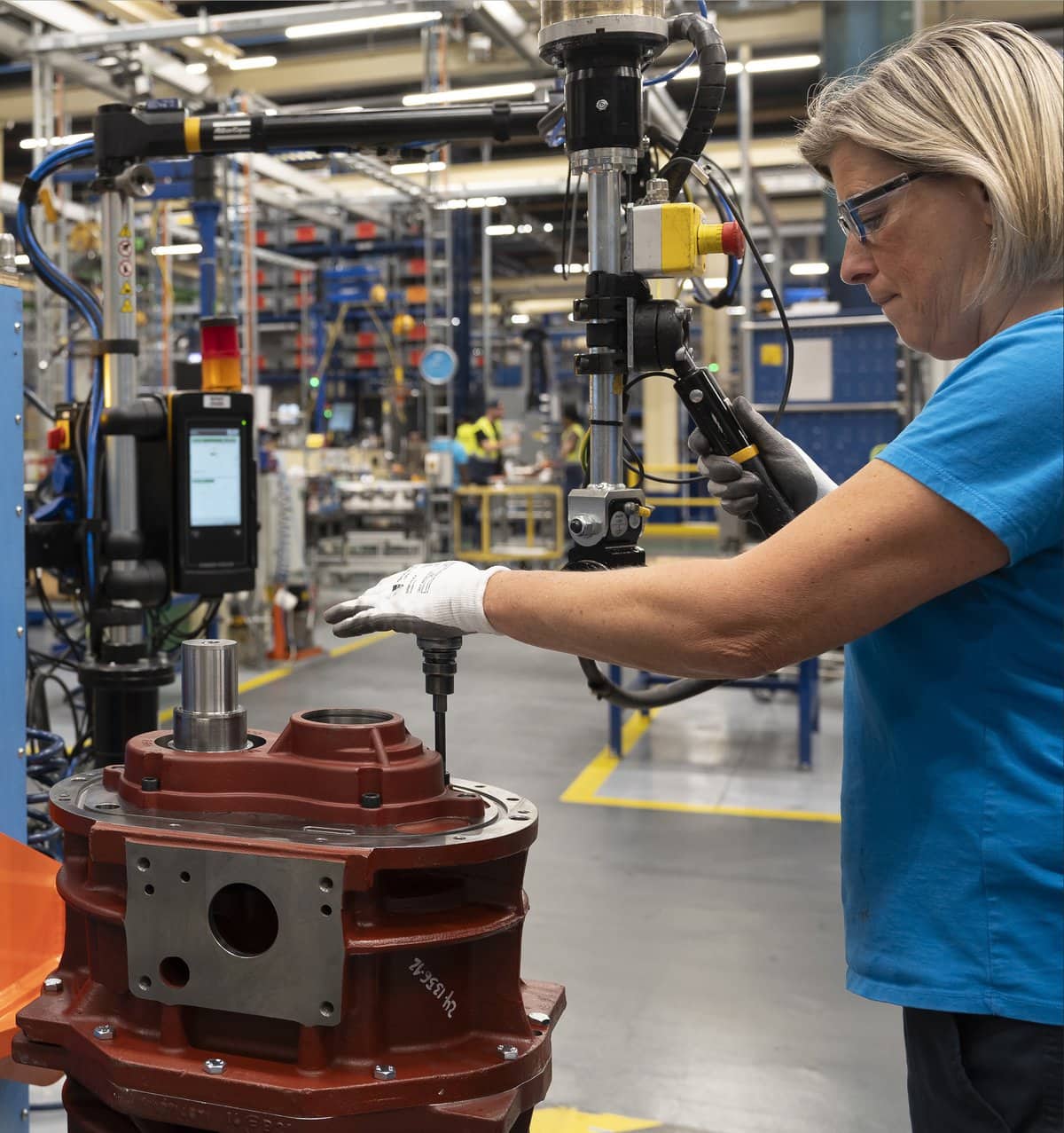 The image shows a woman working in an industrial environment, operating a mechanical tool to assemble a large red metal component. She is wearing safety glasses, a blue shirt, and white protective gloves. The tool appears to be a torque wrench attached to a larger system, which is being used to tighten bolts on the component.