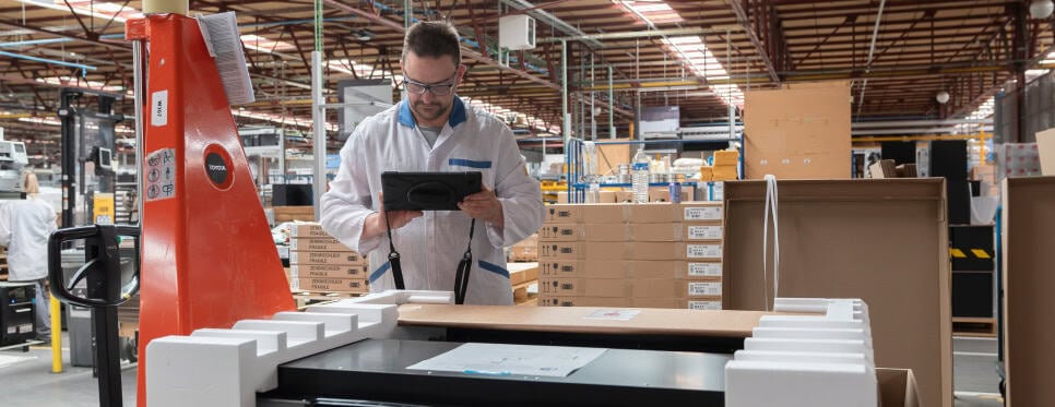 A worker wearing a white lab coat and protective glasses is using a tablet while standing next to a large crate in a warehouse. The background shows rows of shelves filled with boxes and industrial equipment, indicating a busy and organized workspace.