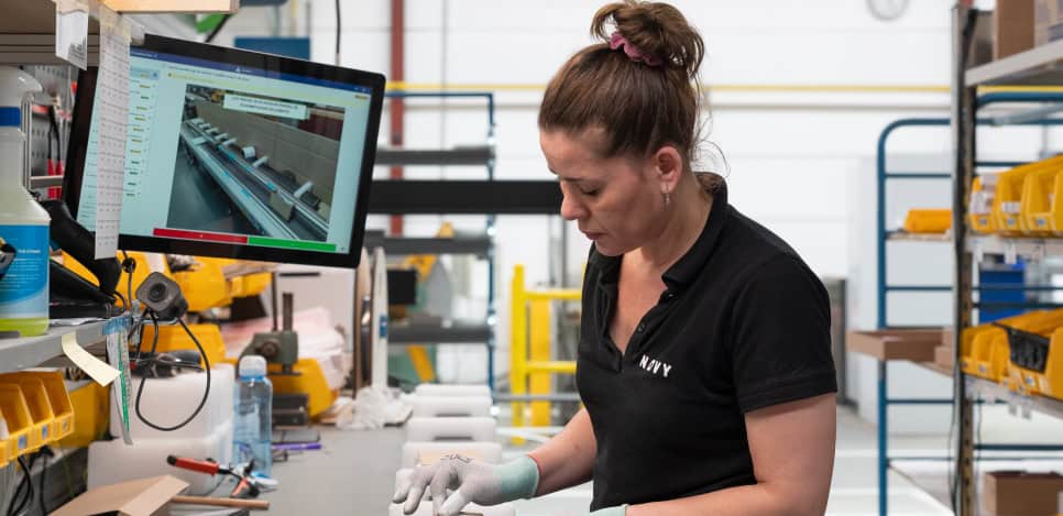 A woman in a black polo shirt works at a cluttered desk in a warehouse. She is handling small objects with gloves. A computer screen displaying a technical diagram is in the background, and shelves with yellow bins surround the workspace.