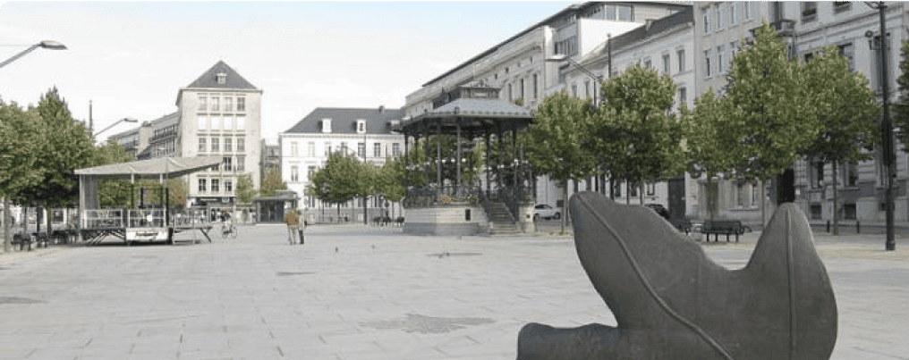 A spacious, paved square is bordered by trees and buildings. In the background, there's a gazebo, and to the right, a modern art sculpture. The square appears empty with only a few people in the distance.