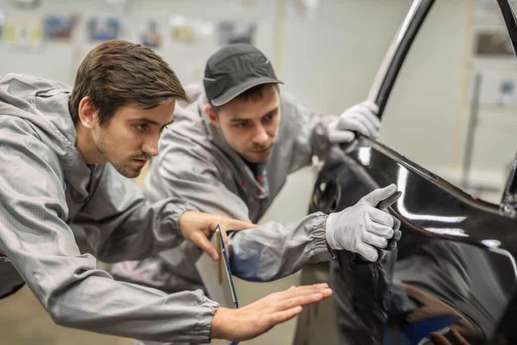 Two workers in gray coveralls and gloves inspect the surface of a car door in a workshop. One holds a tool while the other carefully examines the door, focusing on the quality of the finish. The background is slightly blurred with various equipment and materials.