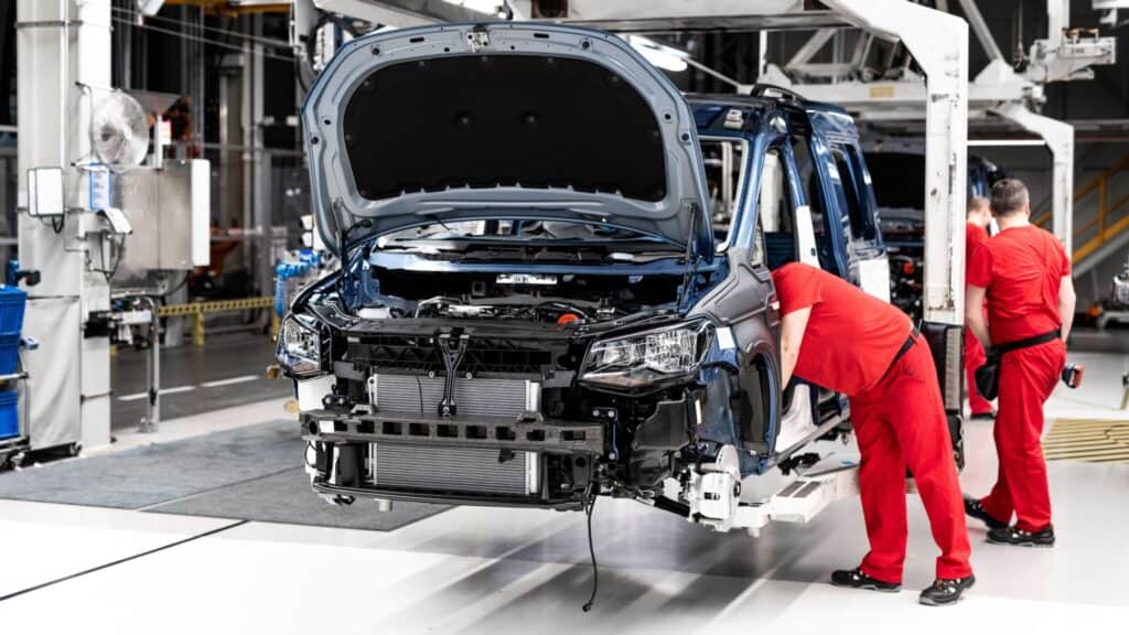 Two workers in red uniforms assemble a partially constructed vehicle on an assembly line in a modern automotive manufacturing facility. The vehicle's front hood is open, exposing its inner mechanical components. The factory setting is organized with machinery visible in the background.