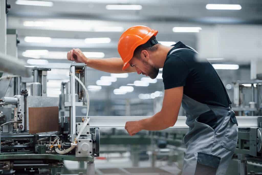 A worker wearing an orange hard hat and gray overalls operates a machine in a factory. He is focused on adjusting a component, with various mechanical parts and equipment visible around him. Bright lights illuminate the industrial setting.