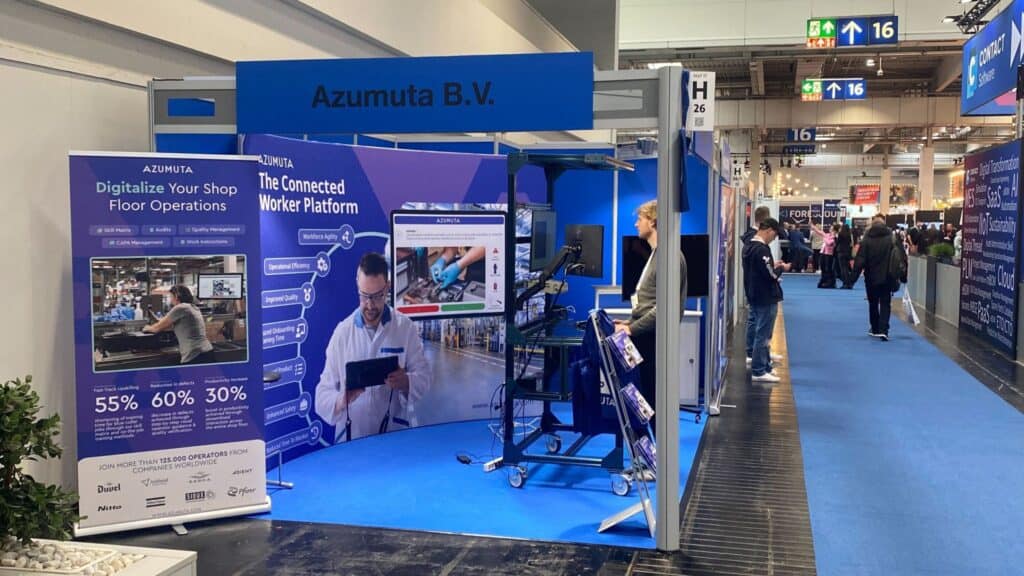 Booth at a trade show with a banner that reads "Azumuta B.V." promoting "The Connected Worker Platform." The display includes a large screen, informational posters, and representatives engaging with visitors. The booth highlights digital shop floor operations and efficiency data.