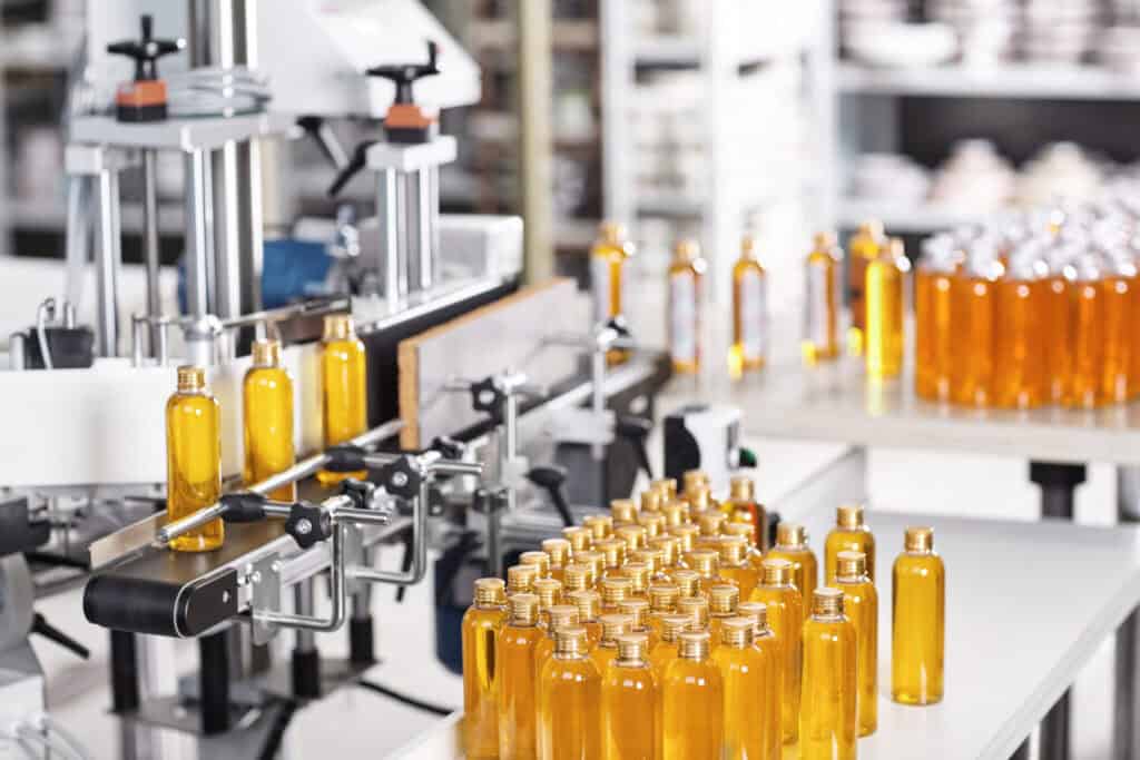 A production line in a factory processes numerous transparent amber-colored bottles filled with liquid. The bottles are aligned on conveyor belts and tables, with machinery and equipment visible in the background. The setting is clean and well-organized.