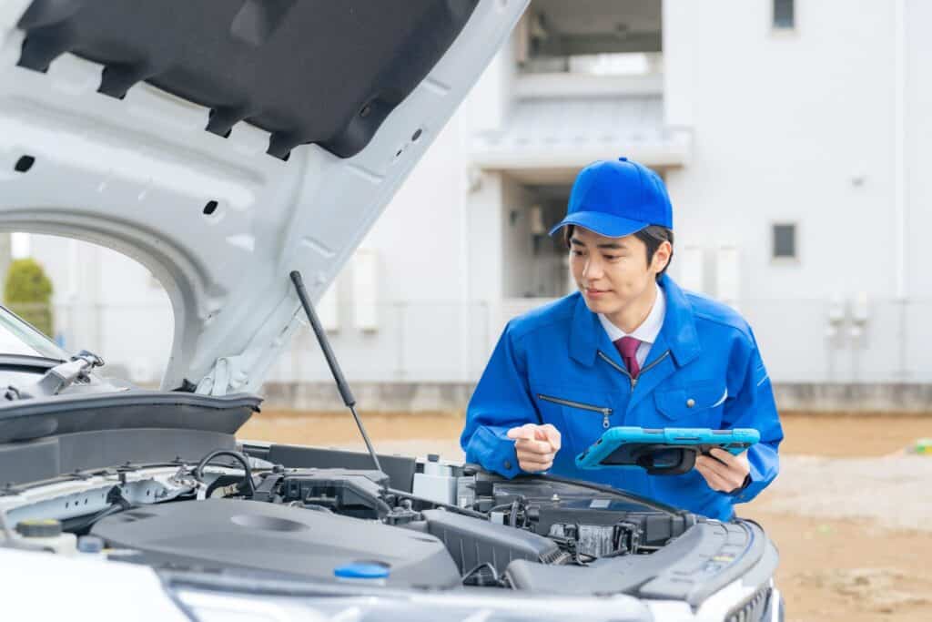 A mechanic dressed in a blue uniform and cap inspects the engine of a car with its hood open. He is holding a tablet and looking intently at the engine, with a partially blurred building in the background.