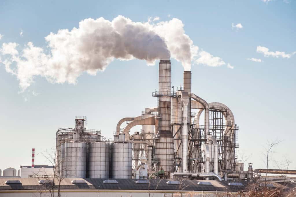 Industrial plant with multiple large chimneys emitting white smoke against a clear blue sky. The plant consists of numerous pipes, tanks, and metal structures. The background sky is free of clouds, highlighting the smoke from the chimneys.