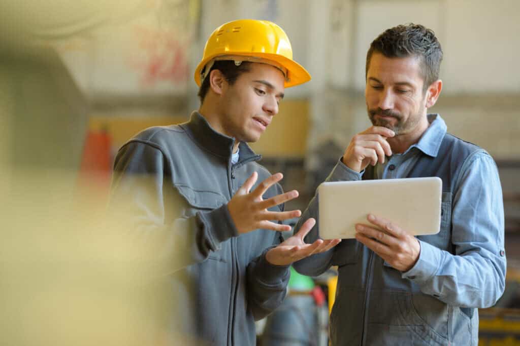 Two male workers stand in a workshop, engaged in a discussion. One, wearing a yellow hard hat, gestures with his hands while explaining something. The other, holding a tablet, listens intently with a thoughtful expression. Both are in work uniforms.