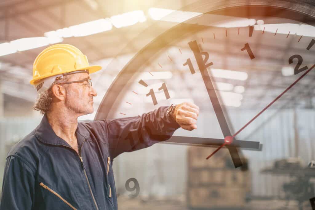 A man with a yellow hard hat and glasses, wearing a blue work uniform, stands in an industrial setting and looks at his wristwatch. A transparent, large clock overlay is superimposed on the image, showing the time as 11:12. Sunlight filters into the background.