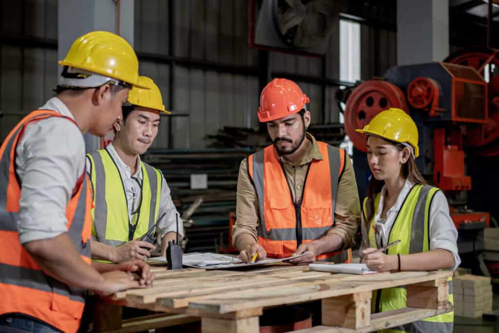 A group of construction workers wearing hard hats and safety vests are gathered around a table. One worker is holding a pen and explaining something on a document, while the others listen attentively. The setting appears to be an industrial or construction site.