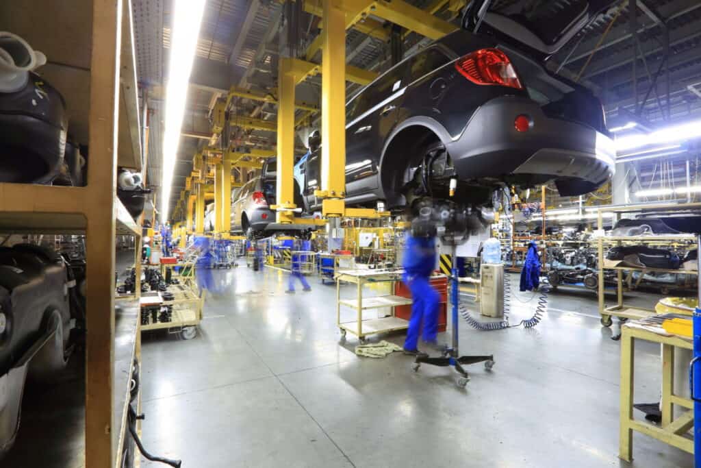 Automobile assembly line with workers wearing blue uniforms. Cars are elevated for assembly and maintenance. The manufacturing facility is well-lit, with a variety of tools and components organized around the workspace.