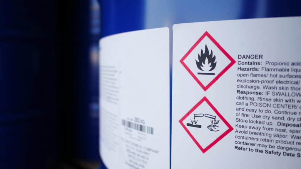 Close-up of a blue chemical drum with warning labels. The labels have hazard symbols, including a flame and a hand being corroded by liquid, and text indicating the substance is highly flammable and corrosive.