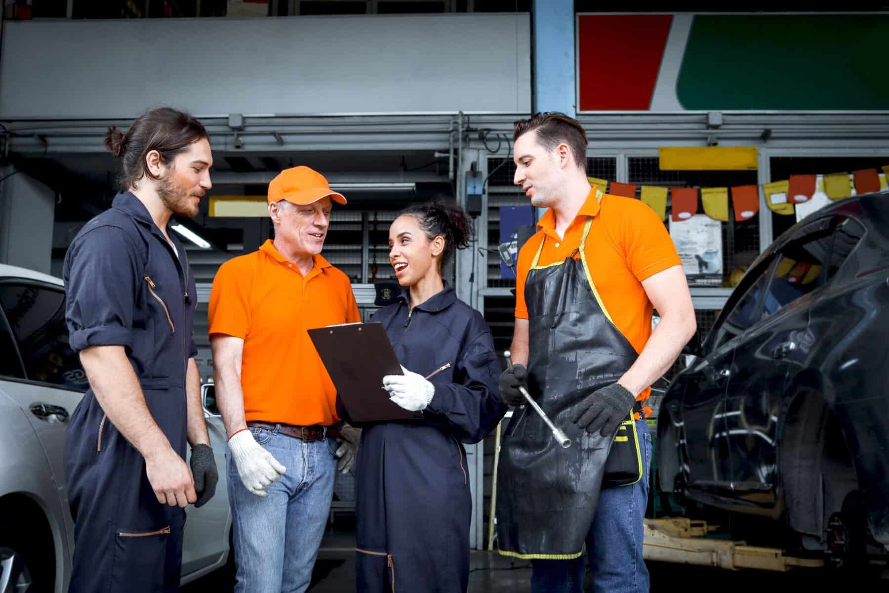 Four automotive technicians, three in dark coveralls and one in an orange shirt and apron, are standing together in a garage. Two are holding tools, while a woman holds a clipboard, and they appear to be discussing something. Cars and workshop equipment are visible in the background.
