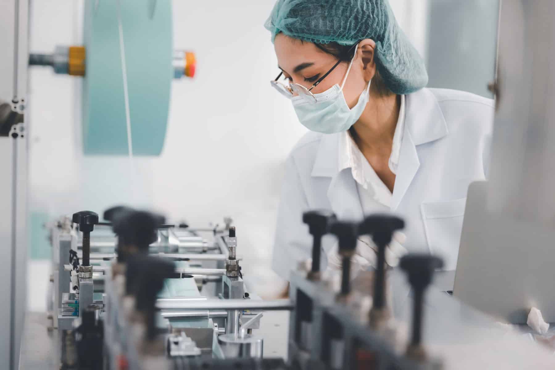 A person wearing a white lab coat, hairnet, glasses, and a face mask is working in a manufacturing or laboratory setting. They are closely inspecting or operating a piece of machinery, focusing intently on their task.