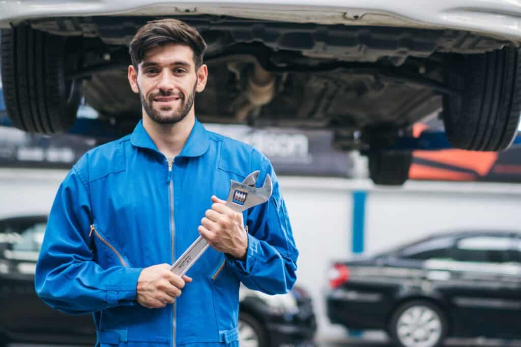 A mechanic in a blue jumpsuit holds a large wrench and smiles at the camera. He stands under a lifted car in a garage, with two parked cars visible in the background.