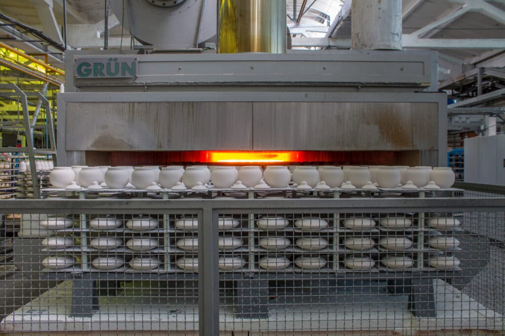 Porcelain cups being fired in a ceramic kiln at an industrial facility. Rows of cups are visible entering the kiln, with a bright orange glow emanating from the interior, indicating high temperatures. The surrounding area appears to be part of a large factory.
