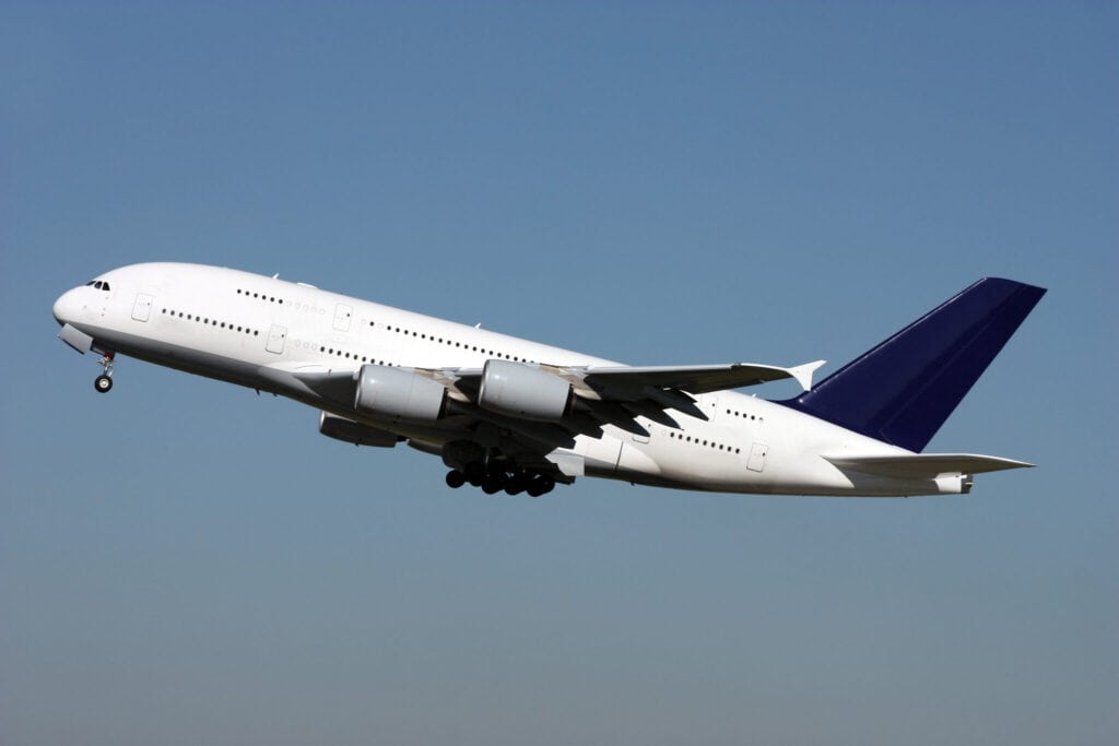 A large, white commercial airplane with a dark blue tail is taking off against a clear blue sky. The landing gear is partially retracted as the plane begins its ascent.