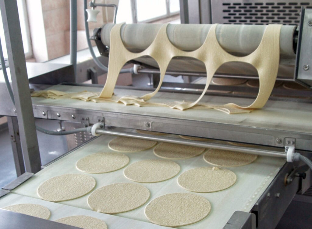 An industrial machine producing large sheets of dough, cutting out circular shapes. The cut dough circles are arranged on a conveyor belt, while the leftover dough scraps are visible on the upper roller. The setup appears to be in a commercial kitchen.