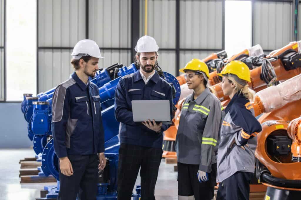 Four workers wearing safety gear, including helmets and reflective vests, stand in an industrial setting. They are gathered around a laptop, which one person is holding, while robotic machinery is visible in the background.