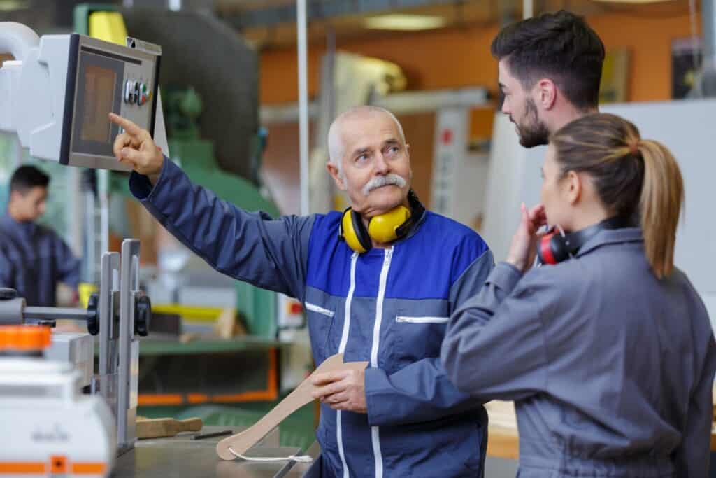 An older man wearing a blue work jacket and yellow ear protection instructs two younger adults in a woodshop. The man is pointing at a machine panel, while the younger adults, dressed in gray overalls with ear protection, look on attentively.