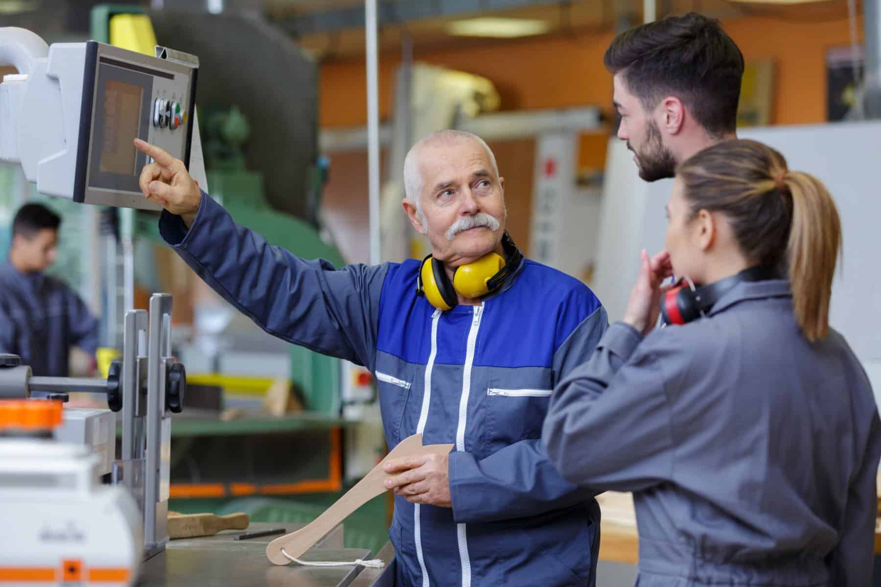 An older man wearing a blue work jacket and yellow ear protection instructs two younger adults in a woodshop. The man is pointing at a machine panel, while the younger adults, dressed in gray overalls with ear protection, look on attentively.