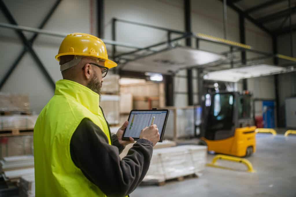 A worker in a yellow hard hat and safety vest uses a tablet in a warehouse. In the background, there is a forklift and stacks of materials. The environment is industrial, with high ceilings and metal beams.