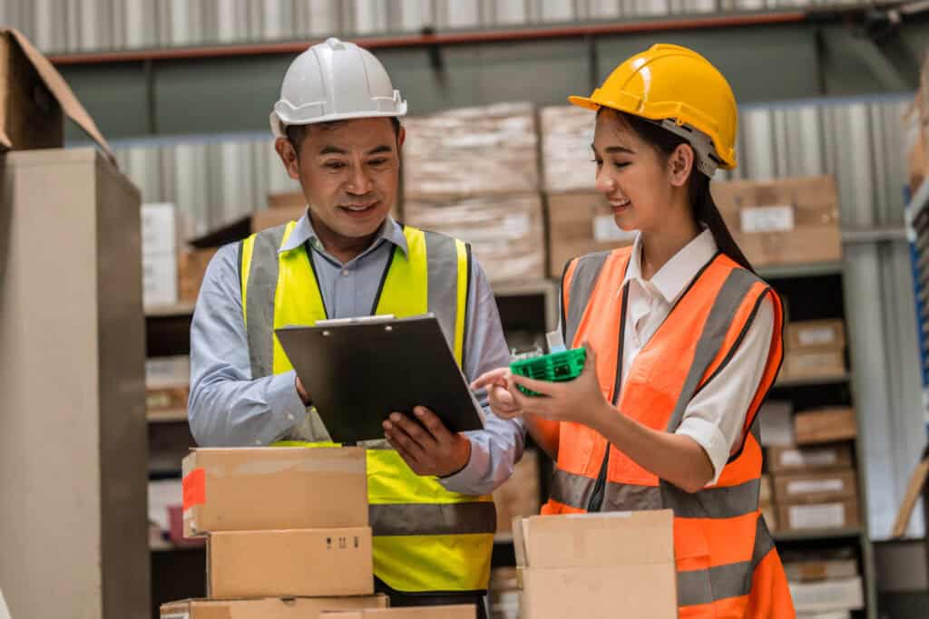 Two workers are in a warehouse, both wearing safety vests and hard hats. The man on the left is holding a clipboard and smiling, while the woman on the right is showing him a small green tool and smiling. In the background, there are shelves stacked with boxes.
