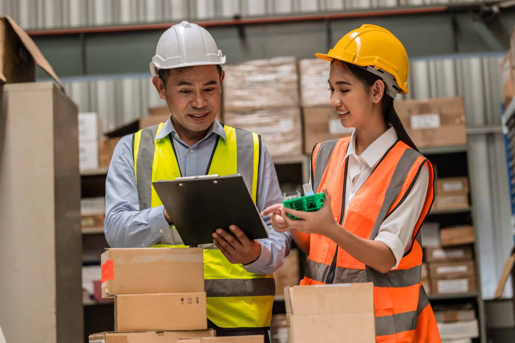 Two workers are in a warehouse, both wearing safety vests and hard hats. The man on the left is holding a clipboard and smiling, while the woman on the right is showing him a small green tool and smiling. In the background, there are shelves stacked with boxes.