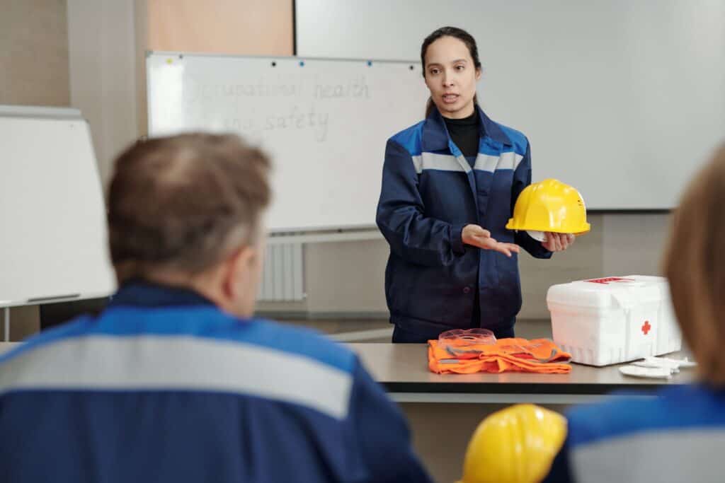 A woman in a blue safety uniform holds a yellow hard hat while giving a presentation to a group. She stands in front of a whiteboard with notes on occupational health and safety. Safety gear, including a first aid kit, is displayed on the table beside her.