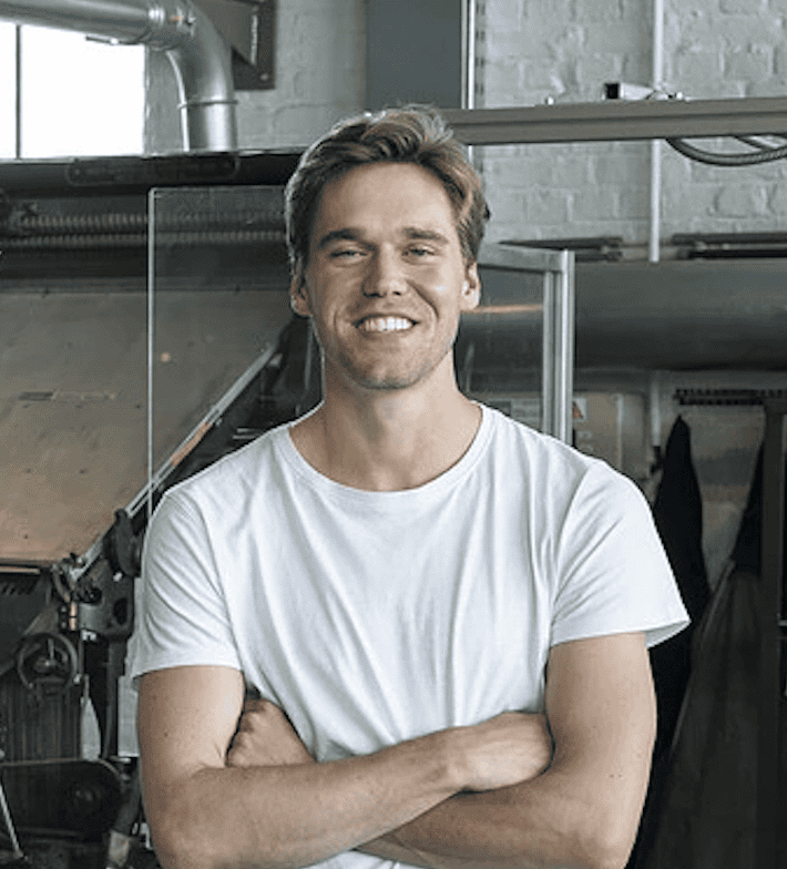 A man with light brown hair, wearing a white t-shirt, stands in front of industrial machinery with his arms crossed. He is smiling and appears to be in a workshop or factory setting with various equipment and tools around.