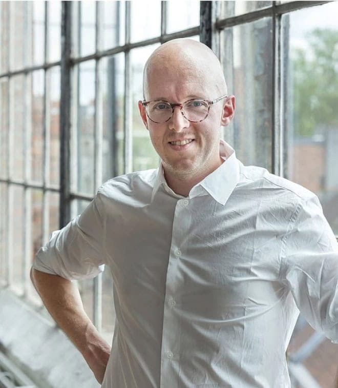 Azumuta's CEO, Batist Leman, standing in front of large industrial-style windows. He is wearing a white shirt and is smiling at the camera, with one hand on his hip. The background shows a blurred view of buildings and greenery outside the windows.