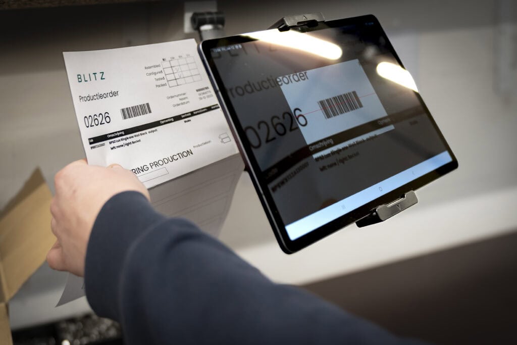 The image shows a person scanning a production order document using a tablet mounted on a stand. The tablet screen displays a magnified view of the document with a barcode highlighted, indicating the use of a scanning application.
