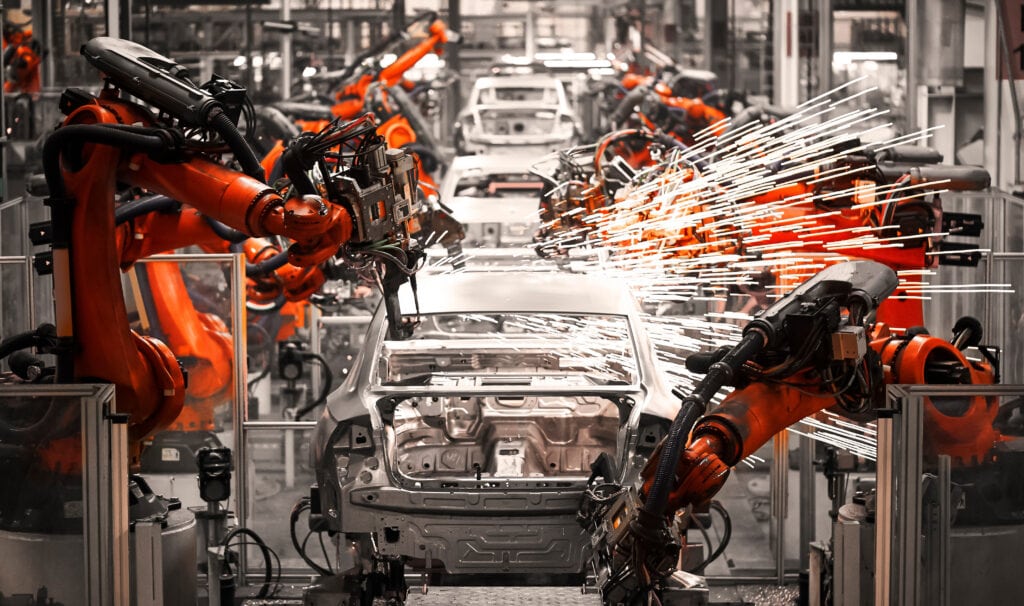 Automated manufacturing assembly line with robotic arms welding car frames, creating sparks during production in an industrial facility.