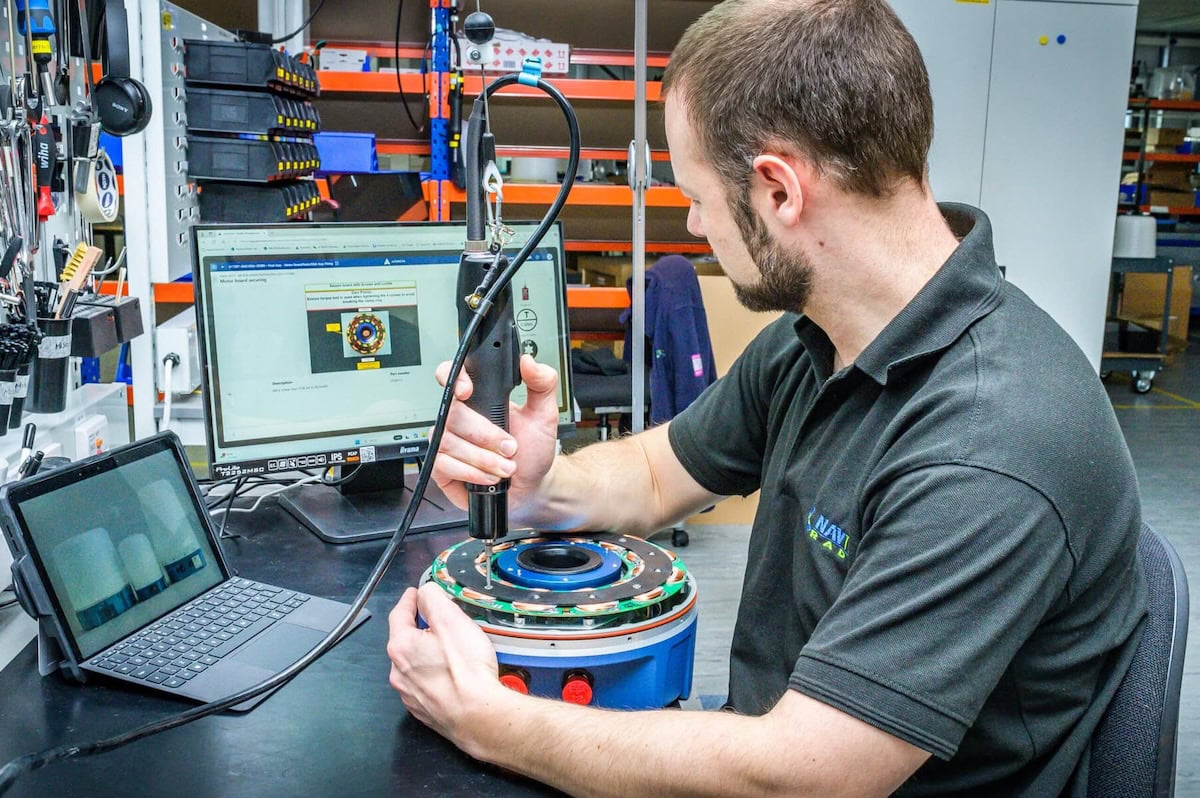 A technician at Navtech Radar is assembling or inspecting a radar component at a workbench. He is using an electric torque tool while following digital work instructions displayed on a monitor. A tablet is also visible beside him, possibly for additional reference or documentation. The workspace is organized with tools hanging nearby and shelves stocked with components in the background.