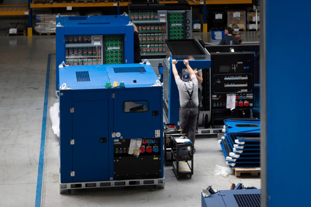 Operators assembling large blue hybrid power units on the shop floor at e-power’s production facility. Visible battery modules and control panels are integrated into the units, with tools and documentation on mobile carts nearby. The environment is organized and structured, highlighting a continuous flow assembly setup.