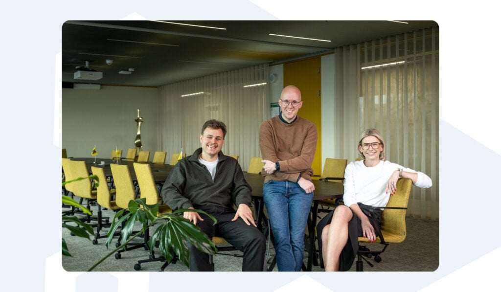 Three Azumuta team members sit and stand together in a bright meeting room with yellow chairs and a modern design. They are smiling at the camera, conveying a friendly and professional atmosphere.