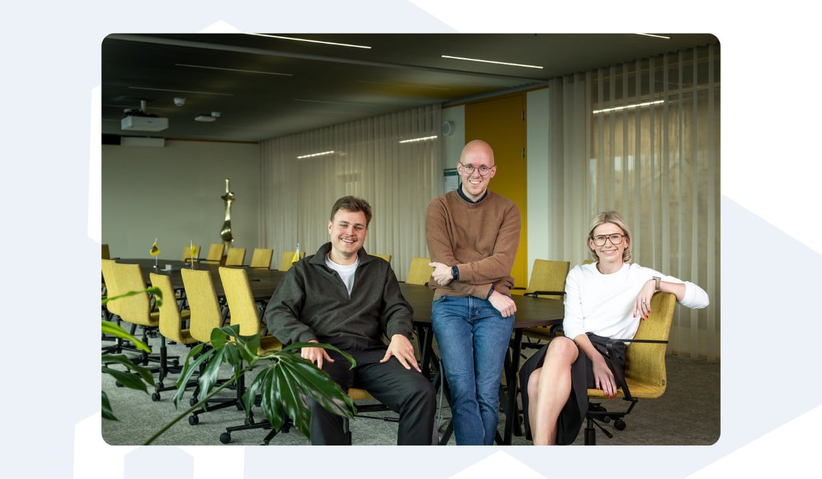 Three Azumuta team members sit and stand together in a bright meeting room with yellow chairs and a modern design. They are smiling at the camera, conveying a friendly and professional atmosphere.